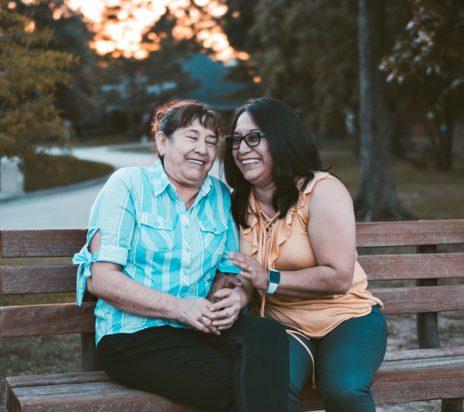 two hispanic women sitting on a bench leaning in towards each other and laughing