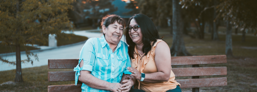two hispanic women sitting on a bench leaning in towards each other and laughing