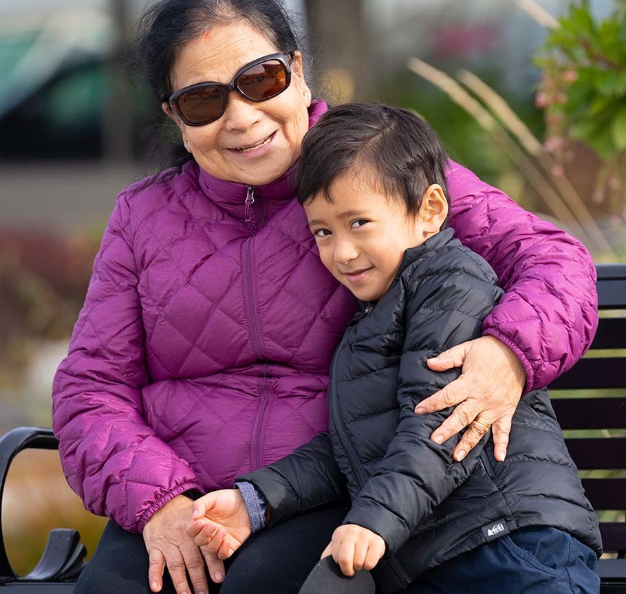 grandmother in sunglasses and magenta down coat sitting on park bench hugs smiling grandson