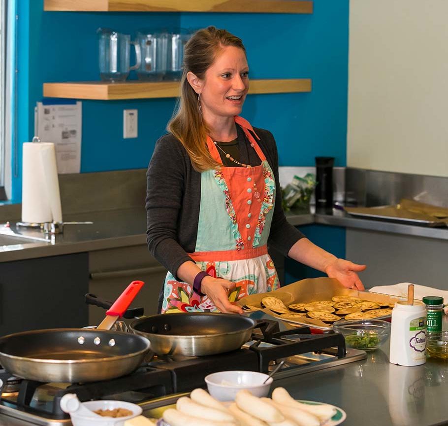 woman in bright orange and blue apron leads cooking class in test kitchen