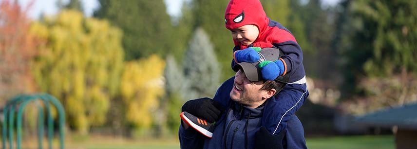 father in baseball cap carries boy in Spiderman costume on his shoulders