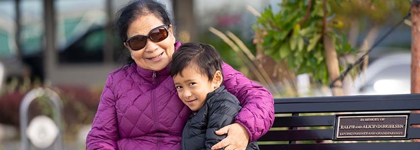 grandmother in sunglasses and magenta down coat sitting on park bench hugs smiling grandson
