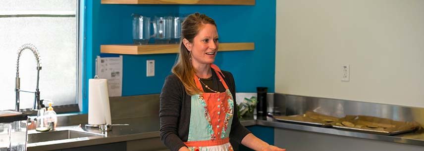 woman in bright orange and blue apron leads cooking class in test kitchen