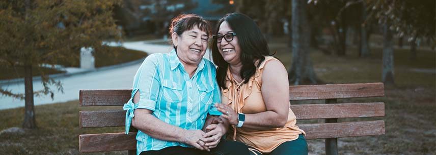 two hispanic women sitting on a bench leaning in towards each other and laughing